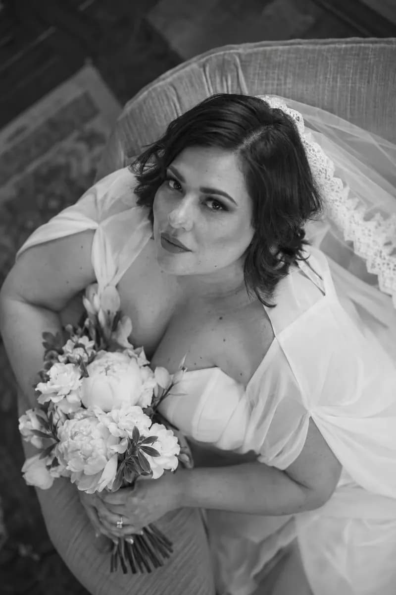 Ilustración creada con IA: novia recostada en un sillón mirando hacia arriba a cámara con el ramo cerca del pecho