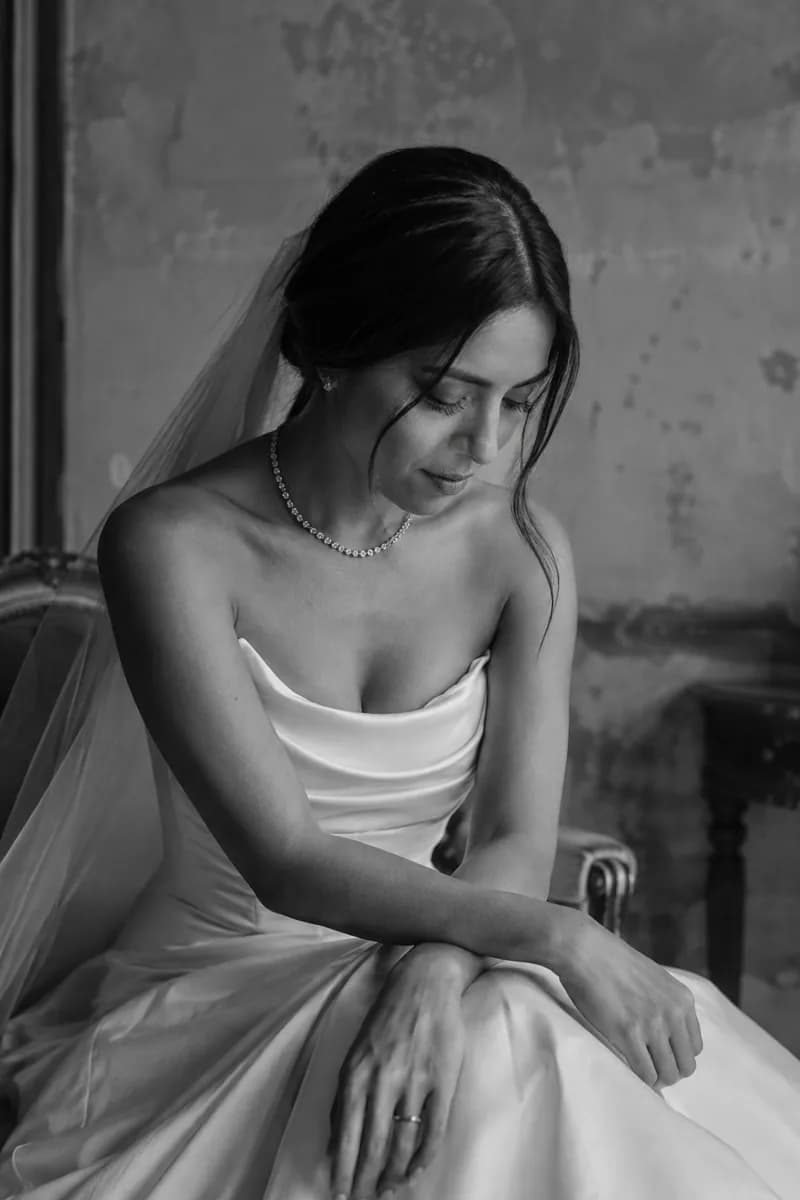 Ilustración creada con IA: novia sentada en tres cuartos con los antebrazos cruzados sobre la falda y mirada baja