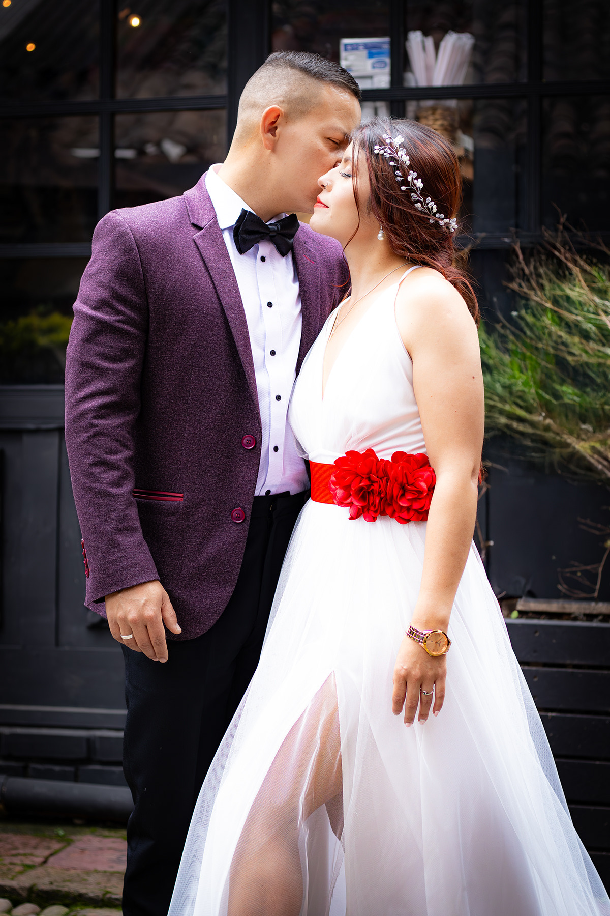 Novio con saco oscuro y corbatín besando la frente de la novia frente a una puerta de vidrio